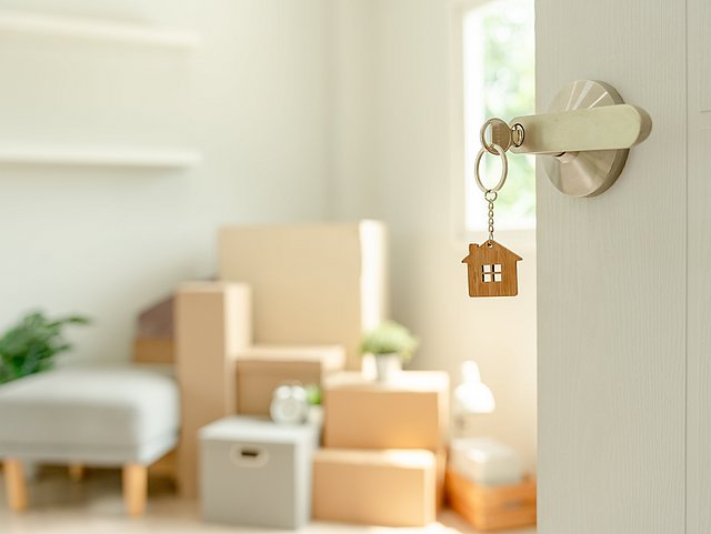 Offene Tür zu einer Wohnung mit Umzugskartons. Ein Schlüssel steckt im Schloss einer Eingangstür. Die Tür ist leicht geöffnet und man erhält einen Einblick in die Wohnung. Neben einigen Kleinmöbeln sind dort allen voran Umzugskartons und Kisten zu sehen.