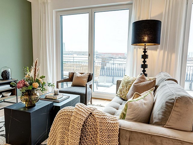 Wohnzimmer Beispiel Einladender Wohnbereich mit gemütlicher Couch mit übergeworfener Decke. Zwei Abstelltische in Granitoptik mit dekorativem Blumenarrangement und einer TV-Ablage. Der Blick führt nach draußen auf den möblierten Balkon.